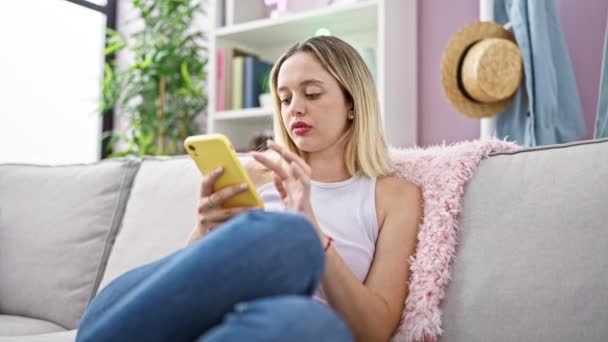 自宅で考えているソファーに座っているスマートフォンを使用して金髪の若い女性 - 映像、動画