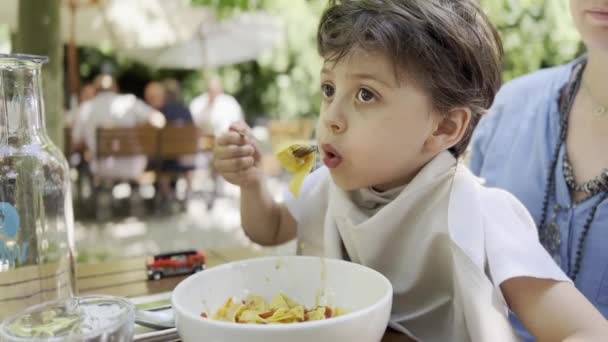 Kid se sienta en la vuelta de mamá en el restaurante al aire libre, enfriando la pasta caliente italiana tradicional - Imágenes, Vídeo