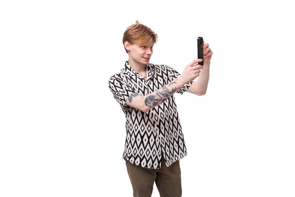 Ένας νεαρός κοκκινομάλλης με καλοκαιρινό πουκάμισο και διαμαντένιο αποτύπωμα βγάζει φωτογραφία στο τηλέφωνο.. - Φωτογραφία, εικόνα