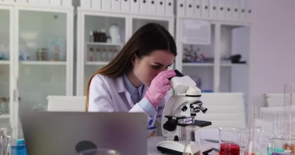 La investigadora examina muestras biológicas bajo el microscopio y escribe los resultados de la investigación en la computadora portátil, en el laboratorio científico. Asistente de laboratorio femenina trabajando en laboratorio - Metraje, vídeo