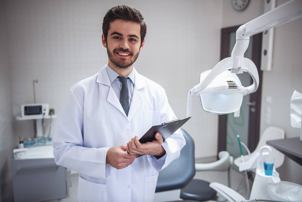 Knappe jonge tandarts in witte jas is een map houden, camera kijken en glimlachen terwijl staande in zijn kantoor - Foto, afbeelding