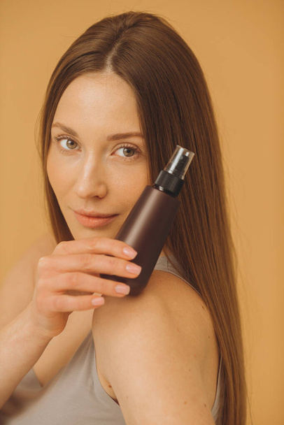 Retrato de mujer hermosa con piel limpia y fresca sosteniendo botella de maquillaje posando sobre fondo beige. - Foto, imagen