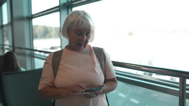Mulher idosa caucasiana esperando a partida no aeroporto em suas férias, usando seu telefone celular  - Filmagem, Vídeo