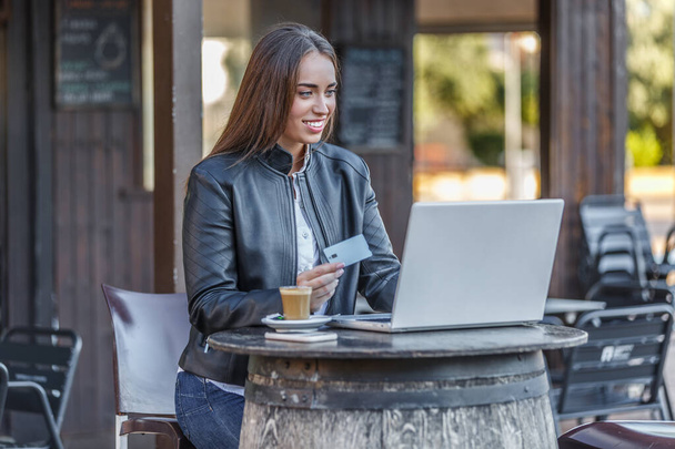 Positieve jonge vrouw in casual outfit zitten aan tafel in de straat cafetaria en browsen netbook terwijl het betalen van online voor de aankoop met plastic kaart - Foto, afbeelding