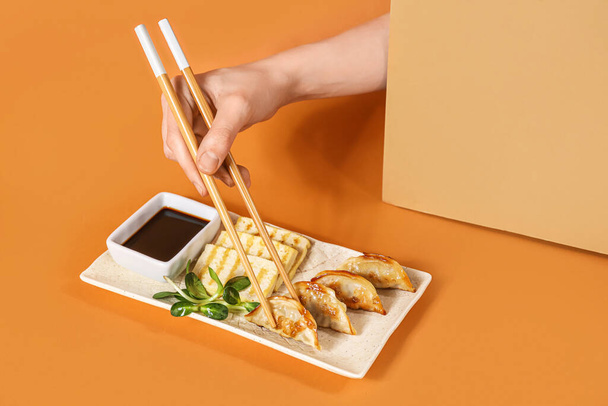 Женская рука с палочками для еды, китайские пельмени, сыр тофу и соевый соус на цветном фоне - Фото, изображение