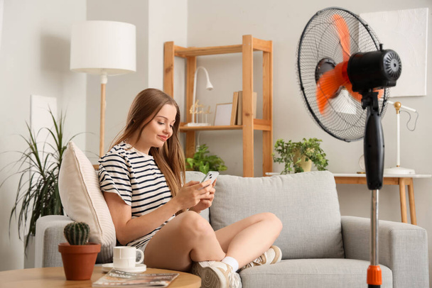 Jeune femme avec soufflage ventilateur électrique en utilisant un téléphone mobile à la maison - Photo, image