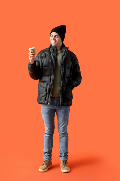 オレンジ色の背景にコーヒーのカップと冬の服の若い男 - 写真・画像