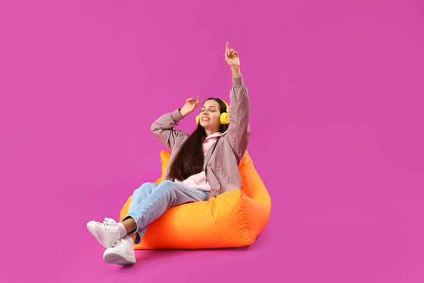 紫色の背景に座っているジャケットとヘッドフォンの若い女性 - 写真・画像