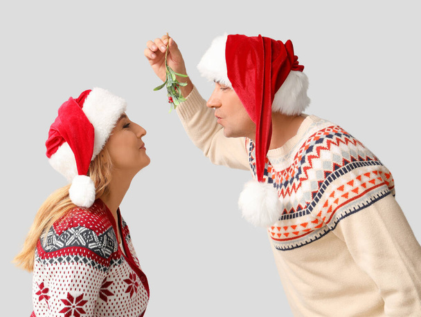 Зрелая пара в шляпах Санты целуется под веткой омелы на светлом фоне - Фото, изображение
