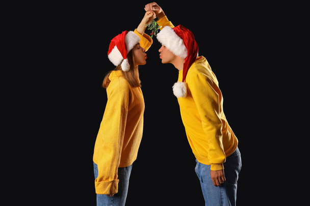 Jeune couple dans Santa chapeaux baisers sous branche de gui sur fond sombre - Photo, image