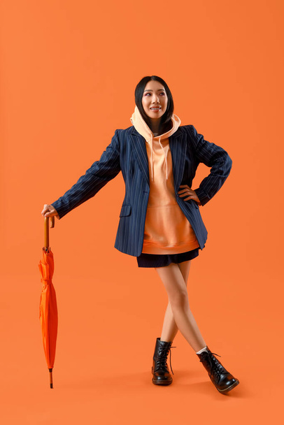 Young Asian woman in hoodie with umbrella on orange background - Photo, Image
