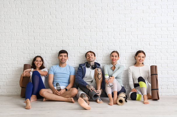 Группа спортивных молодых людей с ковриками для йоги, сидящих возле стены из белого кирпича - Фото, изображение