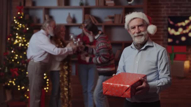 Porträt eines reifen, hübschen Mannes, der ein Weihnachtsgeschenk in der Hand hält und auf die Kamera blickt, während seine Freunde im Hintergrund Spaß haben. Senioren-Neujahrsparty. Winterferienkonzept - Filmmaterial, Video