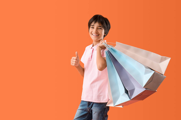 Маленький мальчик с пакетами для покупок показывает большой палец вверх жест на оранжевом фоне - Фото, изображение