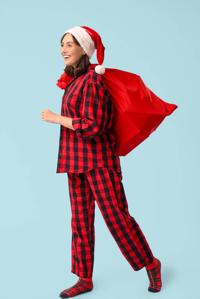 Belle jeune femme en pyjama avec sac Père Noël sur fond bleu - Photo, image