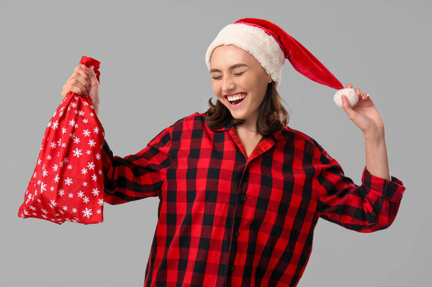 Beautiful young woman in pajamas with Santa Claus bag on grey background - Photo, Image