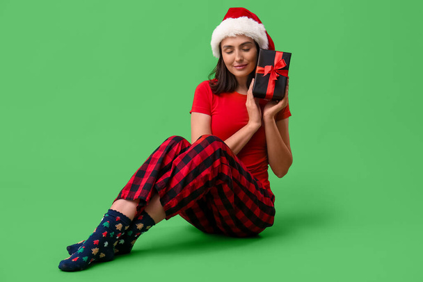 Hermosa mujer joven en pijama y sombrero de Santa con caja de regalo sentado sobre fondo verde - Foto, Imagen