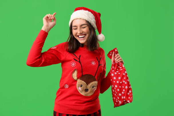 Beautiful young woman in pajamas with Santa Claus bag on green background - Photo, Image