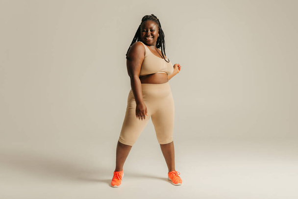 Felice donna africana curvy in abbigliamento sportivo guardando fiducioso nel suo corpo mentre in piedi su sfondo studio - Foto, immagini