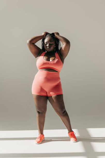 Full length of confident African plus size woman in sportswear adjusting her dreadlocs while standing in fitness studio - Zdjęcie, obraz