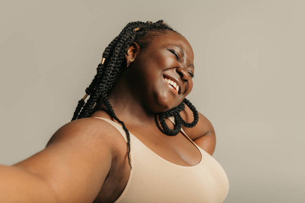 Joyful plus size African woman in sportswear making selfie while standing on studio background - Photo, Image