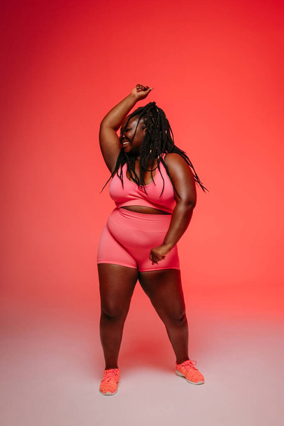 Happy African plus size woman in sportswear dancing and radiating self-love against vibrant background - Fotó, kép