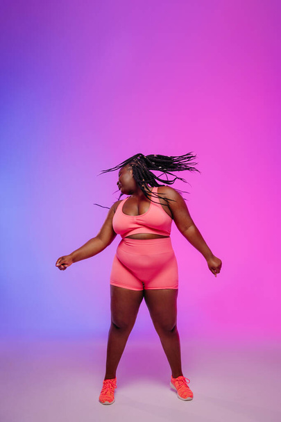 Beautiful African plus size woman in sportswear dancing and shaking dreadlocs against colorful background - Foto, immagini