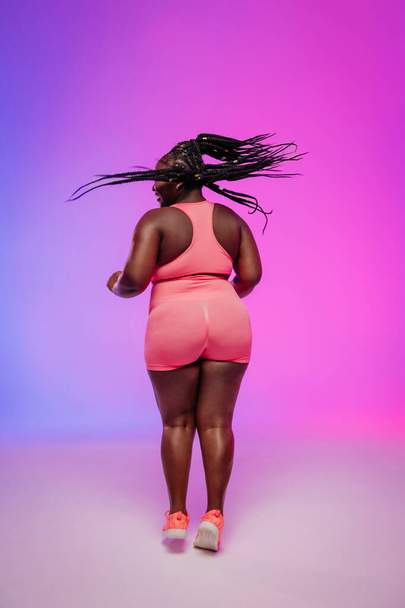 Rear view of African curvy woman in sportswear dancing and looking confident against vibrant background - Фото, изображение