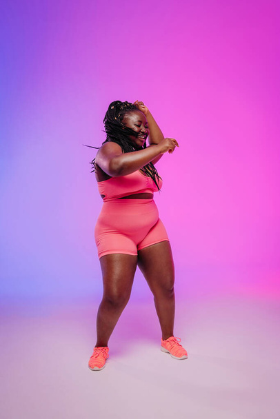 Joyful African curvy woman in sportswear dancing and radiating self-love against vibrant background - Photo, image