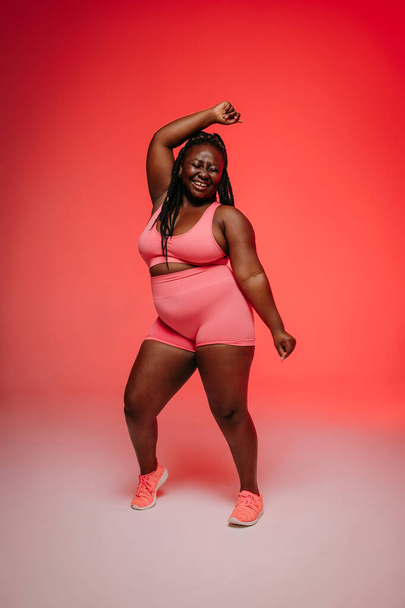 Happy African plus size woman in sportswear dancing and looking confident against vibrant background - Fotó, kép