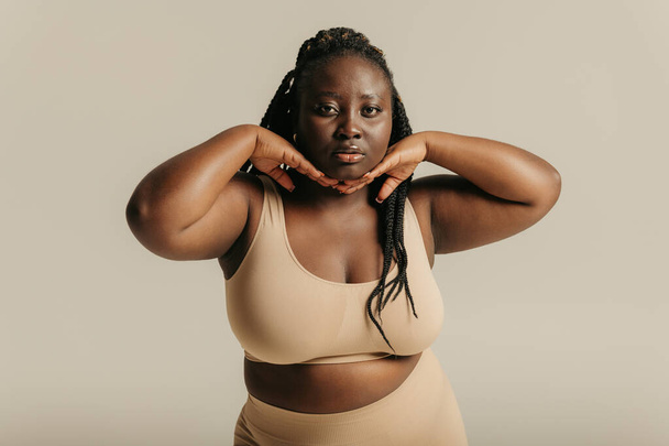 Beautiful curvy African woman in underwear touching face and looking at camera on studio background - 写真・画像