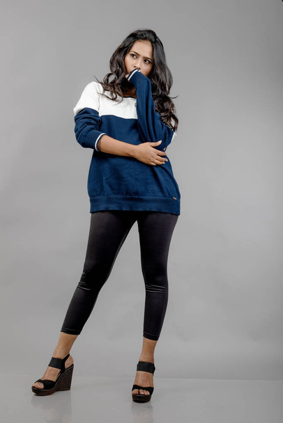 Intialainen nuori naismalli rennoissa talvivaatteissa harmaata taustaa vasten. Pitkä musta tukka malli yllään musta leggingsit, sininen ja valkoinen huppari. - Valokuva, kuva