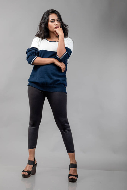 Ινδή νεαρή γυναίκα μοντέλο σε casual winterwear σε γκρι φόντο. Μακριά μαύρα μαλλιά μοντέλο φορώντας μαύρα κολάν, μπλε και άσπρο φούτερ. - Φωτογραφία, εικόνα