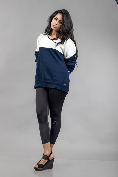 Indiai fiatal női modell alkalmi télikabátban szürke háttérrel. Hosszú fekete hajú modell fekete leggings, kék és fehér pulóver. - Fotó, kép