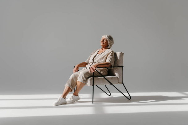 Relaxed gray hair senior woman sitting in comfortable chair and looking calm against background - Фото, зображення