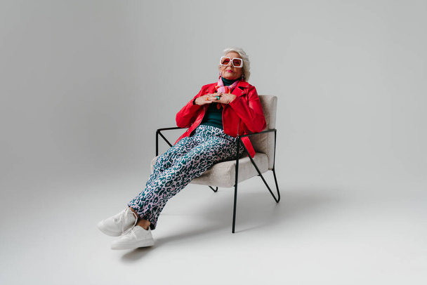 Cool starší žena v módní oblečení sedí v pohodlném křesle na šedém pozadí - Fotografie, Obrázek