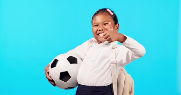 Girl child, soccer ball and studio for thumbs up, backpack or smile on face for sport, learning and blue background. Young female kid, school uniform or football with icon, sign language and portrait. - Footage, Video