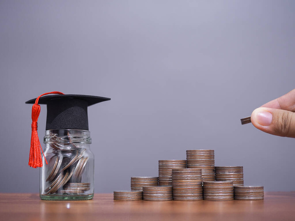 Close up hand putting coin in stack of coins and money in the glass bottle with graduation hat. The concept of saving money for education, student loan, scholarship, tuition fees in future - Photo, Image