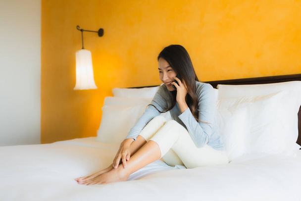 Πορτρέτο όμορφη νεαρή Ασιάτισσα γυναίκα χρησιμοποιώντας κινητό τηλέφωνο στο κρεβάτι με φλιτζάνι καφέ στο εσωτερικό του υπνοδωματίου - Φωτογραφία, εικόνα