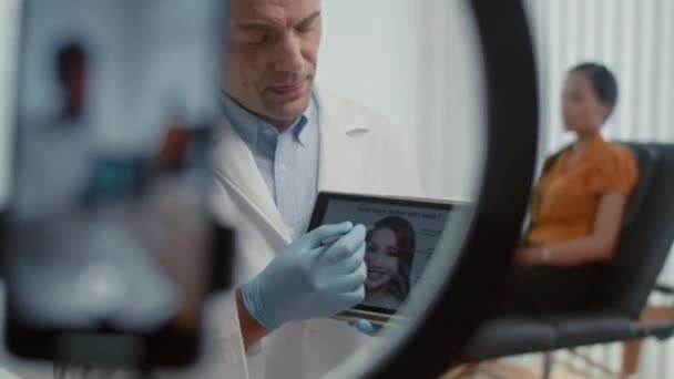 Médico masculino hablando sobre el procedimiento de botox en cámara mientras hace video para su blog usando smartphone, tableta digital y luz de anillo, paciente femenino sentado en segundo plano - Imágenes, Vídeo