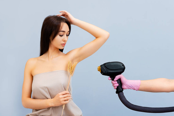 Epilation. A beautiful young woman holds several ears of wheat near her armpit. Cosmetologists hand holds a laser device from the right side. The concept of laser hair removal. - Photo, Image