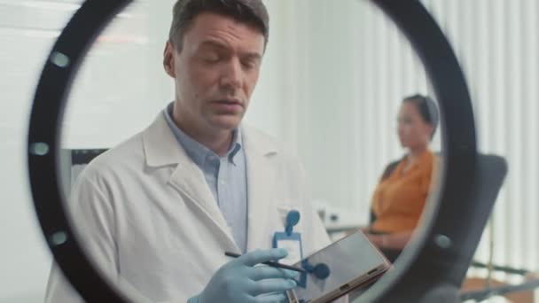 mężczyzna chirurg plastyczny mówi o zabiegu kosmetologicznym podczas strumienia przy użyciu selfie pierścień światła w klinice - Materiał filmowy, wideo