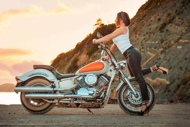 Όμορφη σέξι γυναίκα με ψηλά τακούνια και δερμάτινο παντελόνι, ποζάροντας με αυτοπεποίθηση με μοτοσικλέτα. Ηλιοβασίλεμα στο βάθος. Η έννοια της Ημέρας των μοτοσικλετιστών. - Φωτογραφία, εικόνα