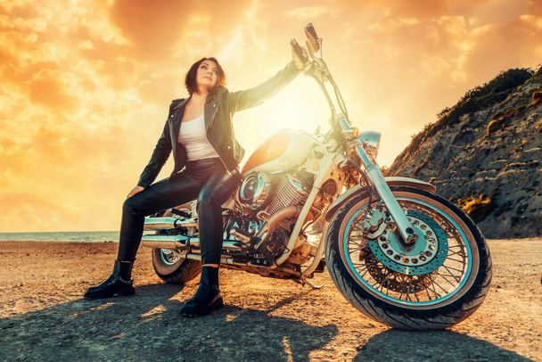 Una hermosa mujer guay en una chaqueta de cuero y pantalones, posando sentado en una motocicleta, sobre el fondo de una puesta de sol épica. Vista de abajo. El concepto de viaje en moto. - Foto, imagen