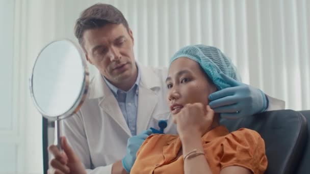 Μέτριο πλάνο του γυναικείου Ασιάτη ασθενή συζητώντας το αποτέλεσμα του λίφτινγκ με πλαστικό χειρουργό στην κλινική - Πλάνα, βίντεο