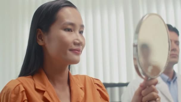 Střední záběr mladé asijské ženy dívá na sebe v zrcadle po facelift procedury provedené lékařem na klinice - Záběry, video