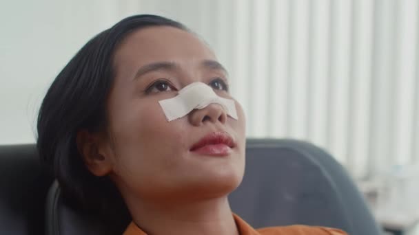 Portret młodej Azjatki z opaską na nosie leżącej na kanapie w gabinecie chirurgii plastycznej i patrzącej w kamerę - Materiał filmowy, wideo