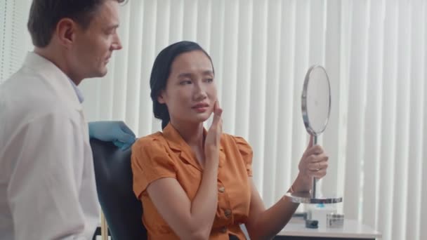 Medium shot of young Asian woman looking at her face in mirror after beauty procedure sitting next to male doctor in clinic - Footage, Video