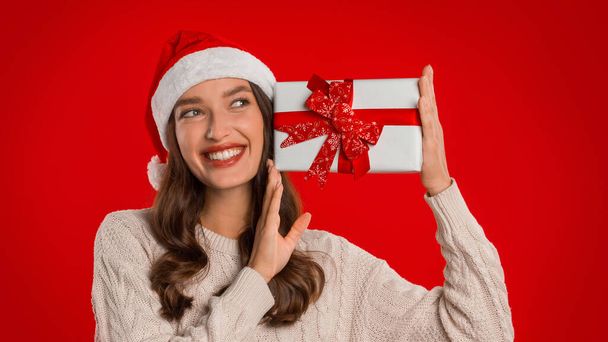Счастливая симпатичная женщина в шляпе санта и обычном свитере, празднующая Новый год или Рождество, держа подарочную коробку рядом с лицом, на красном студийном фоне, панорама. Специальное предложение на зимний сезон - Фото, изображение