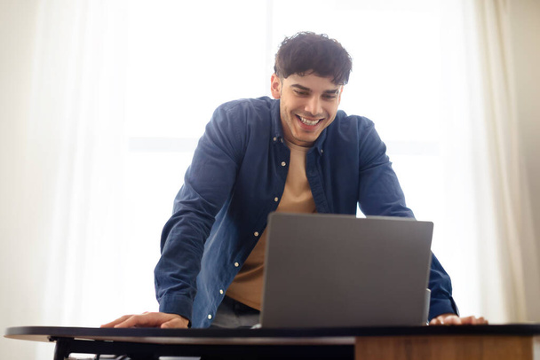 Sonriente hombre guapo de Oriente Medio que trabaja con el ordenador portátil en el lugar de trabajo en el interior de la oficina habitación, de pie cerca del escritorio con el ordenador. Carrera independiente y concepto de negocio online - Foto, imagen